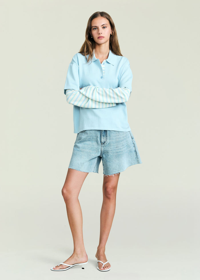 Model wearing Emilee Top in pale blue with a striped underlayer, arms crossed, paired with light wash denim shorts.