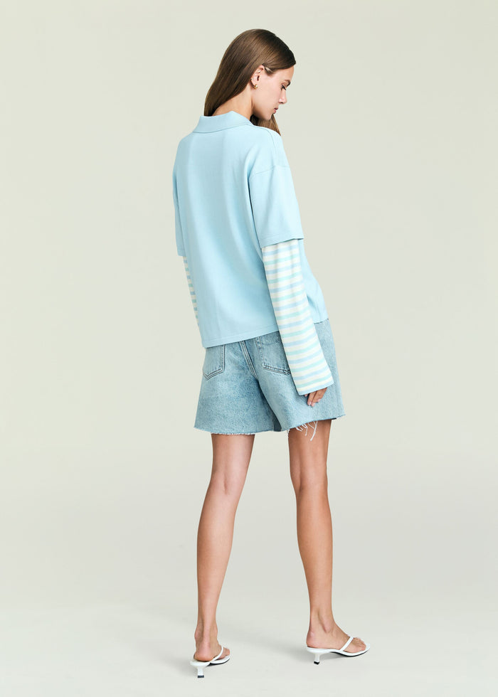 Back view of the Emilee Top in pale blue with a striped long sleeve underneath and denim shorts.