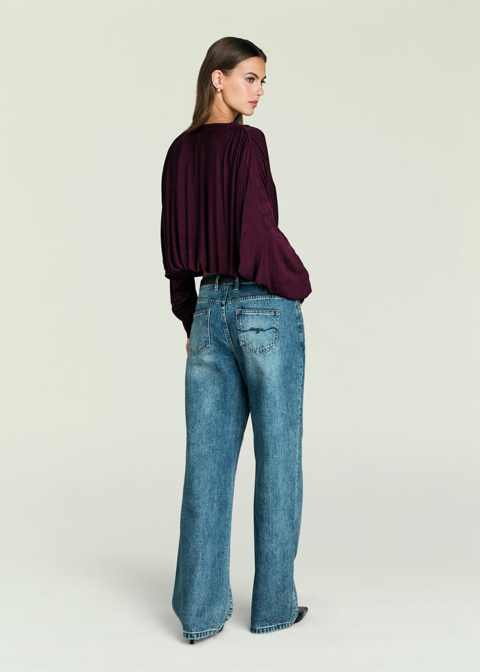 Back view of model wearing burgundy Jerome Top with draped sleeves and jeans.
