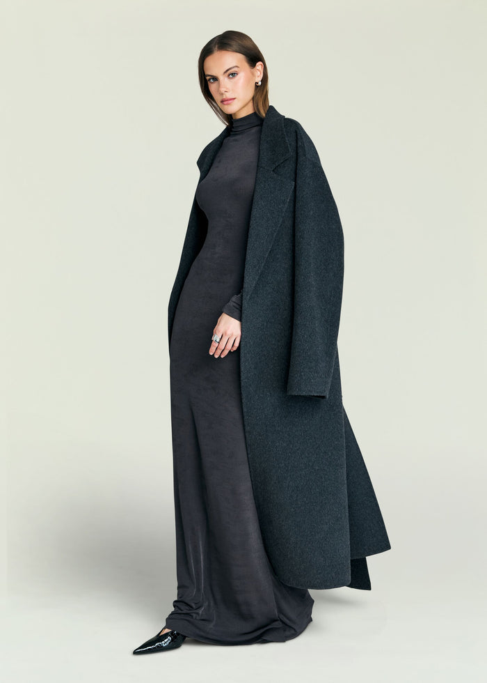 Model wearing a dark charcoal maxi dress with a long overcoat draped over her shoulders.