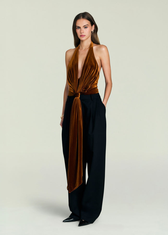 Front view of a model in a copper velvet halter bodysuit with a deep plunging neckline and waist tie, paired with black wide-leg pants.