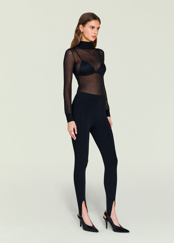 Front view of a model in a black sheer top over a black bra with high waisted stirrup leggings and heels
