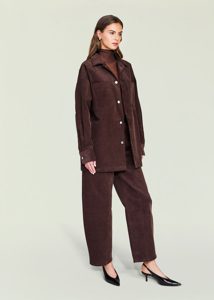 Model wearing an oversized dark brown corduroy shacket with large patch pockets and silver buttons over a brown turtleneck.