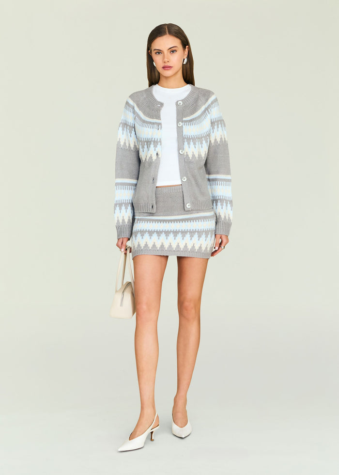 Model wearing a gray blue geometric knit cardigan with a matching mini skirt and holding a beige bag