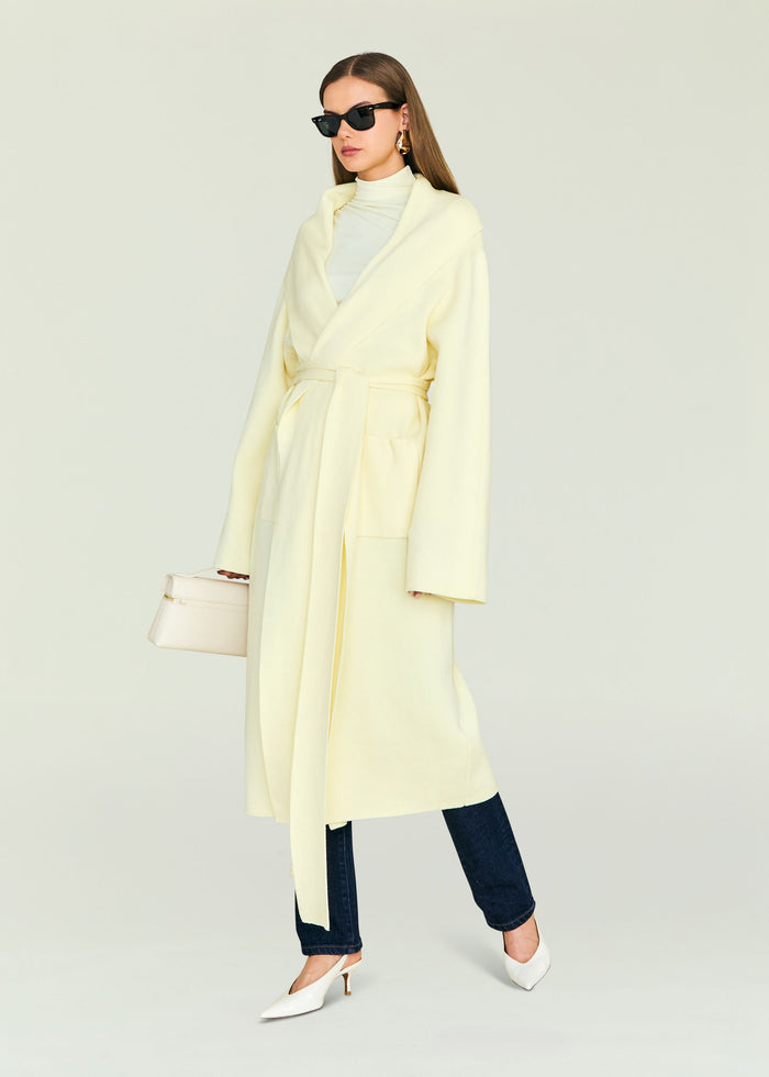 Model wearing pale yellow Crosby Sweater Duster wrap coat with belt, paired with dark jeans and white heels.