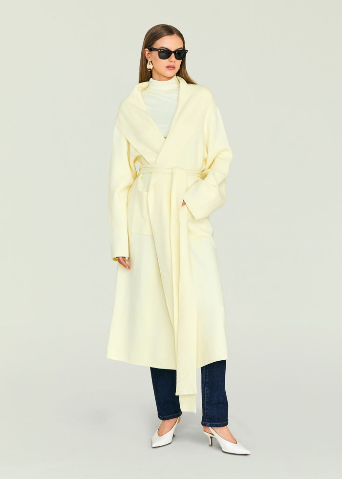 Woman wearing a pale yellow Crosby Sweater Duster coat with a belt, sunglasses, and jeans.