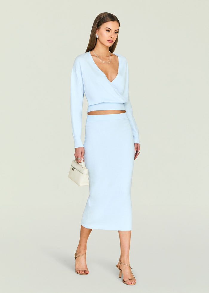 Model wearing a pale blue knit wrap top with a matching midi skirt, accessorized with silver jewelry and a cream handbag.