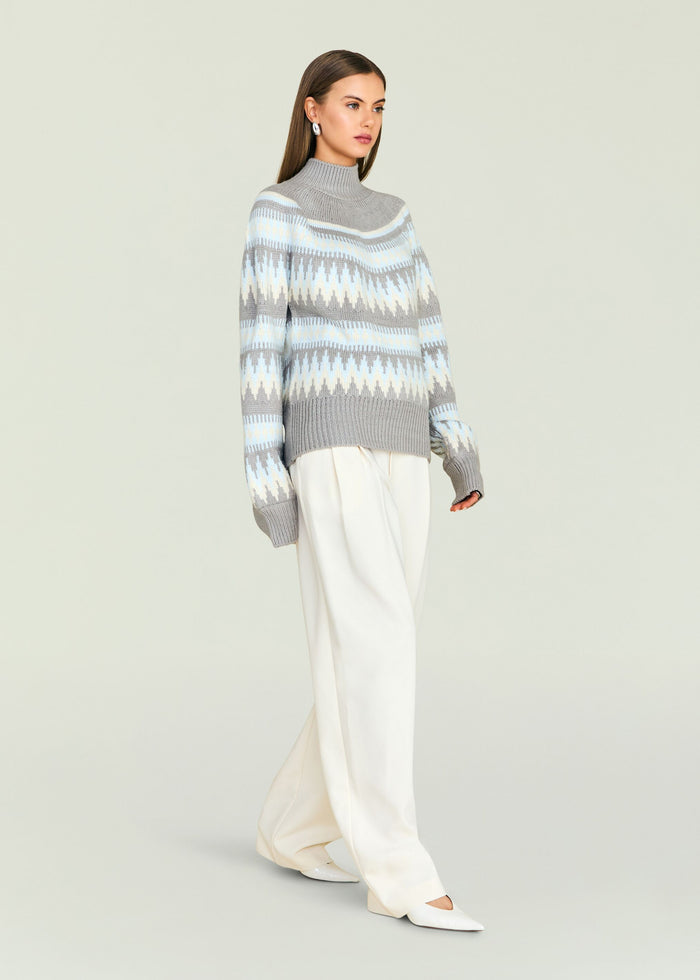 Model in a gray patterned knit turtleneck sweater with light blue and white bands paired with white trousers