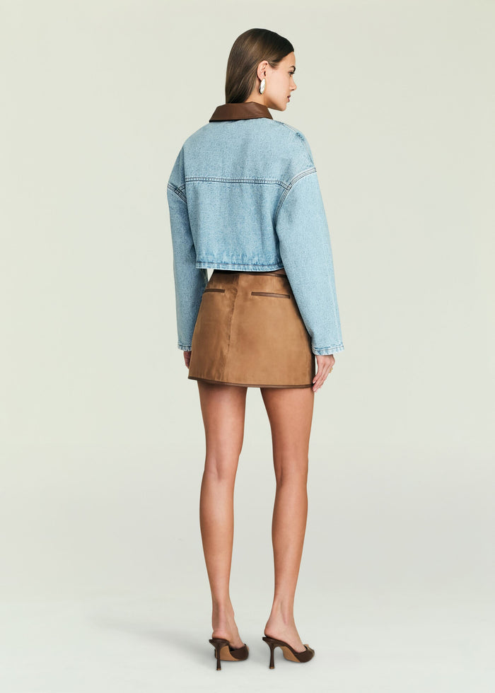 Back view of cropped light denim Coop Jacket with brown leather collar over a white top and tan skirt.