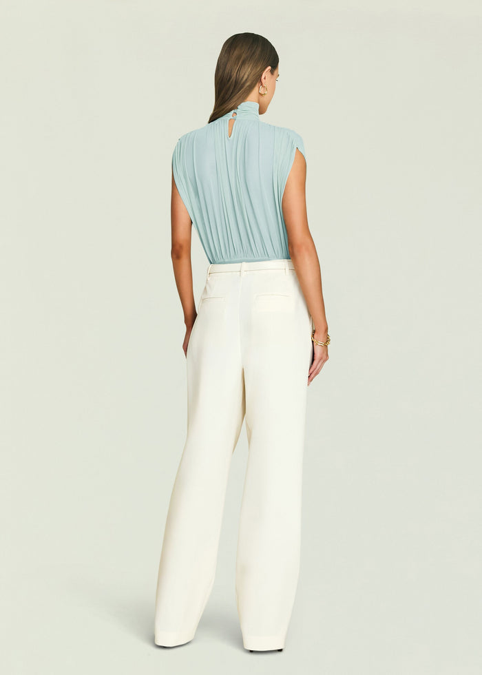 Back view of light blue Giulia bodysuit with pleated draping and a keyhole at the neckline, paired with white trousers.