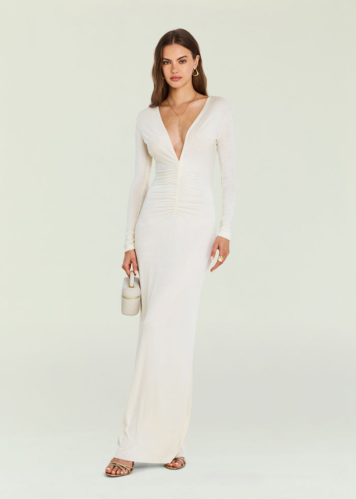 Ebony Maxi Dress worn by a model in ivory with a plunging V neckline long sleeves and ruching at center paired with a small white handbag