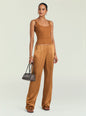 A woman modeling a top and pant in a warm camel brown color, featuring a ribbed knit tank top and wide-leg satin pants, holding a small dark gray handbag against a light gray studio background. thumbnail image 2 of 5