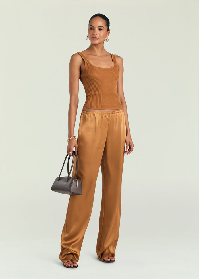 A woman modeling a top and pant in a warm camel brown color, featuring a ribbed knit tank top and wide-leg satin pants, holding a small dark gray handbag against a light gray studio background.