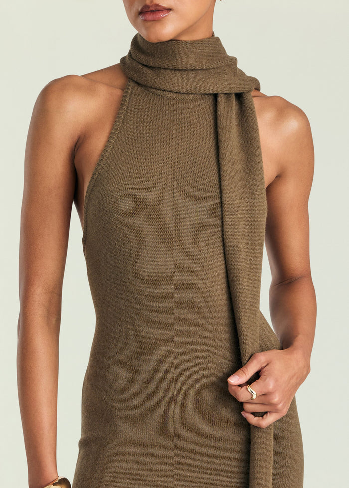 A close-up of a model wearing a ribbed knit halter-neck dress in a warm taupe-brown color, featuring a draped cowl neck detail.