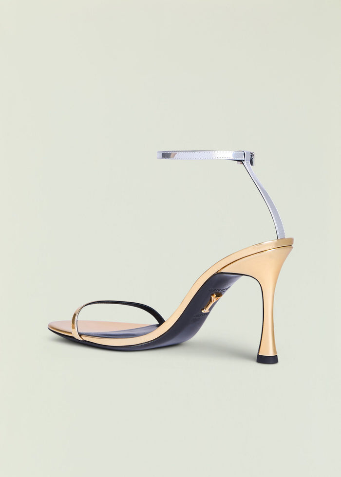 Fowla Sandal featuring a pale beige toe strap and slim metallic ankle strap on a nude stiletto heel