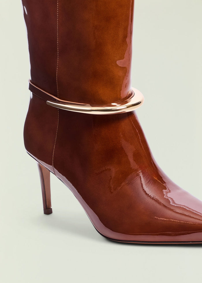 Close up of glossy brown boot shaft showing a curved silver ankle strap detail.