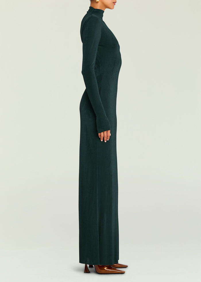 Side view of a model in a dark green velvet Kim Maxi Dress with long sleeves and high neck, paired with brown heels.