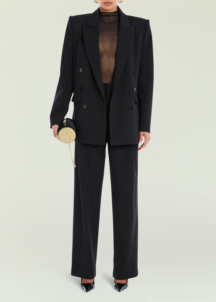 Model in a black tailored double breasted suit with a sheer top, holding a round gold handbag.
