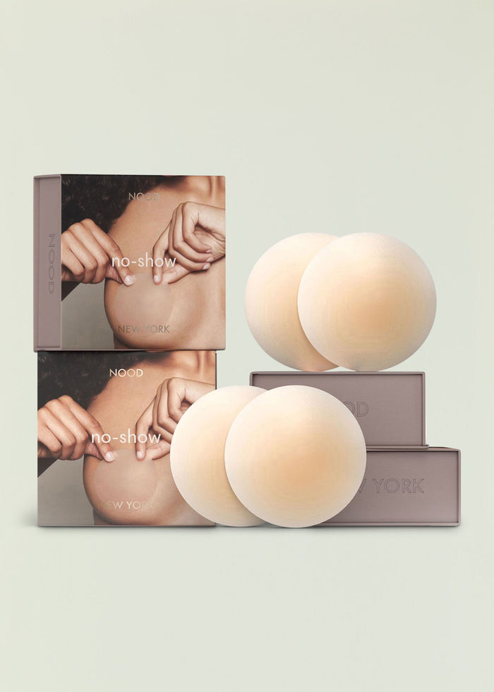 Packaging for no show nipple covers with beige silicone disks displayed beside stacked boxes.