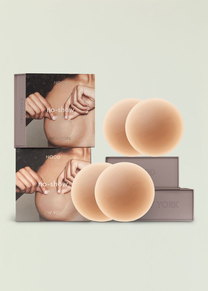 Stacked NOOD packaging beside large nude silicone nipple covers on a pale backdrop.
